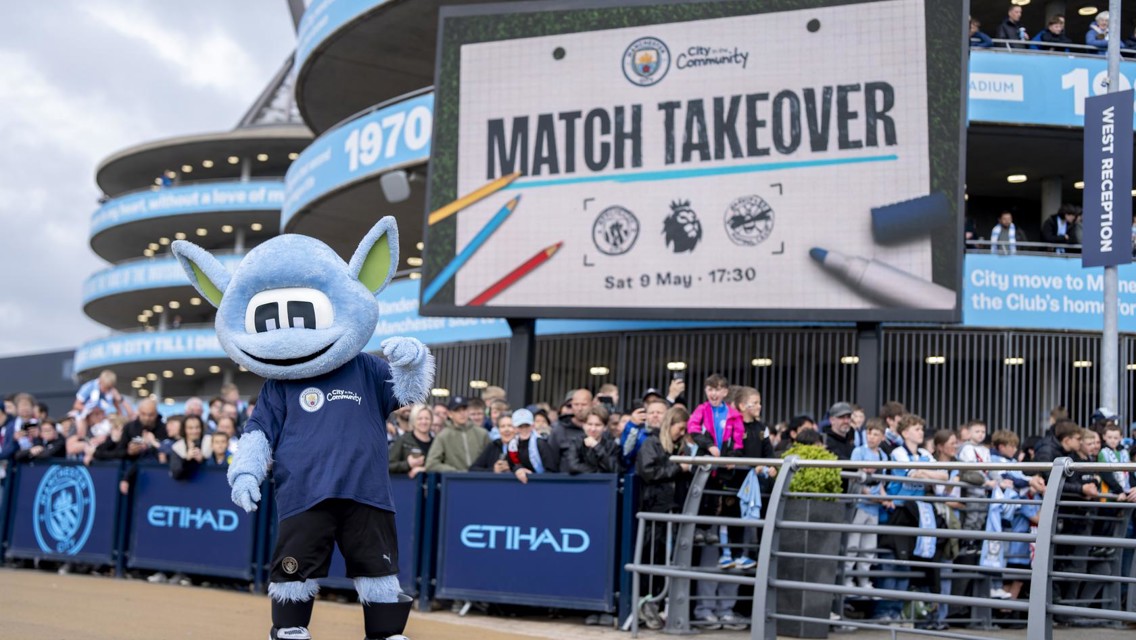 Etihad Stadium with a crowd and a mascot in front of a large sign displaying 'Match Takeover' and the date 'Sat 9 May'.