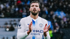 A footballer in a Manchester City uniform is clapping during a match. The player's shirt has the Manchester City logo and the 'No Room for Racism' badge.