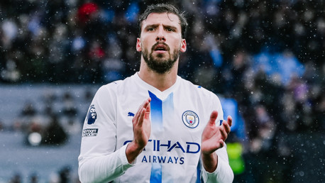 A footballer in a Manchester City uniform is clapping during a match. The player's shirt has the Manchester City logo and the 'No Room for Racism' badge.