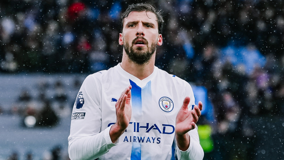 A footballer in a Manchester City uniform is clapping during a match. The player's shirt has the Manchester City logo and the 'No Room for Racism' badge.