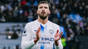 A footballer in a Manchester City uniform is clapping during a match. The player's shirt has the Manchester City logo and the 'No Room for Racism' badge.