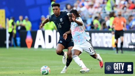 Two soccer players compete for possession of the ball during a match. The player wearing number 11 is in a white and pink uniform, and the other wearing number 16 is in a blue uniform. The match takes place on a grassy field with spectators in the background.