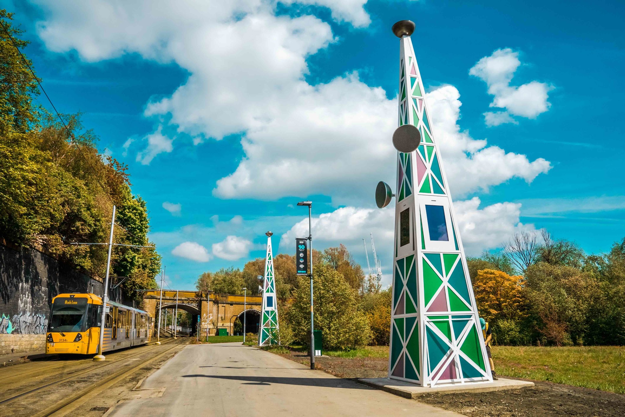 A tram near a station with colorful geometric-patterned towers and graffiti-covered brick walls. An arch bridge is visible in the background. The sky is bright with scattered clouds.
