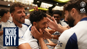 Group of people in Manchester City jerseys celebrating and smiling with hands on each other's heads in a lively setting. Inside City logo and Etisalat logo visible.