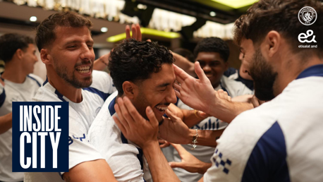 Group of people in Manchester City jerseys celebrating and smiling with hands on each other's heads in a lively setting. Inside City logo and Etisalat logo visible.