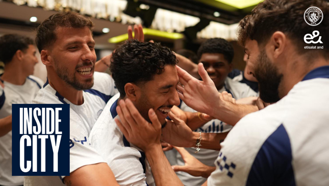 Group of people in Manchester City jerseys celebrating and smiling with hands on each other's heads in a lively setting. Inside City logo and Etisalat logo visible.