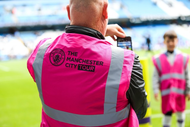 A man wearing a pink high-visibility vest with 'The Manchester City Tour' written on the back, taking a photo with his phone at a football stadium.