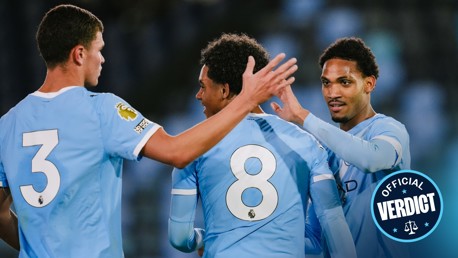 Three Manchester City players in light blue jerseys celebrating during a match with blurred faces.