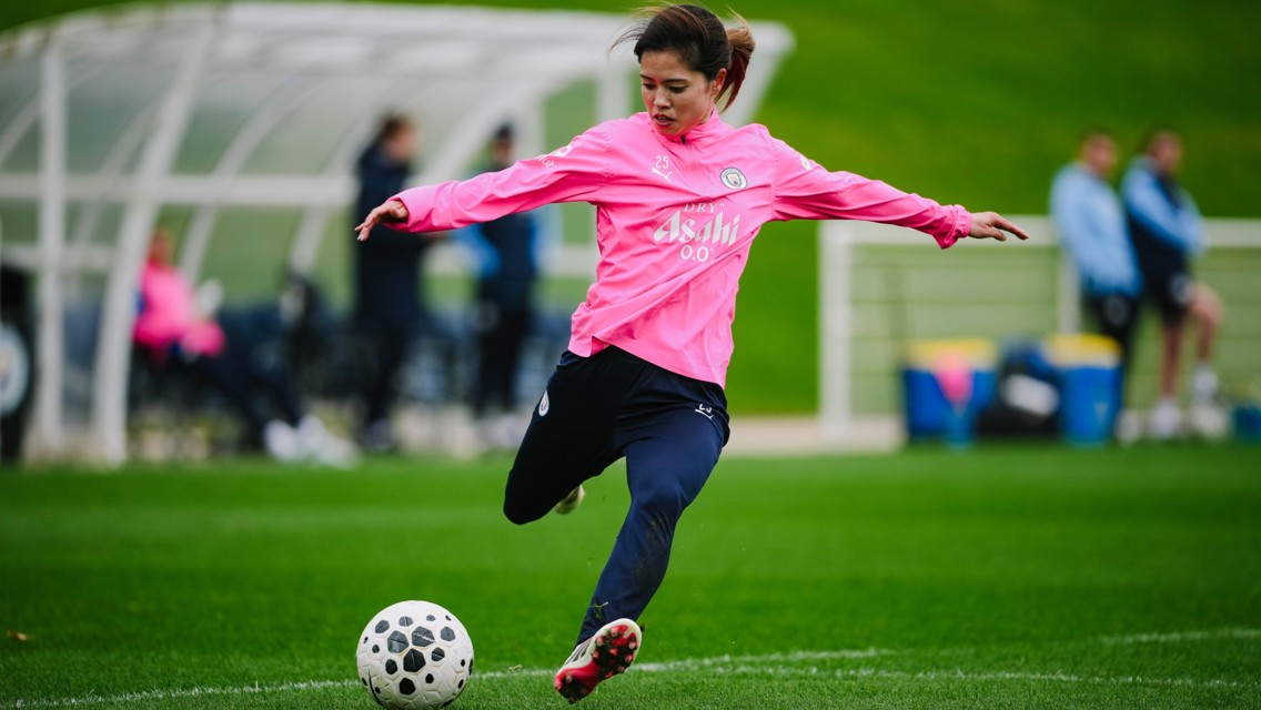 A female football player in a pink jersey is kicking a black and white soccer ball on a green field, with a sideline area in the background.