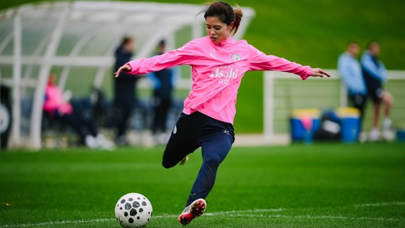 A female football player in a pink jersey is kicking a black and white soccer ball on a green field, with a sideline area in the background.