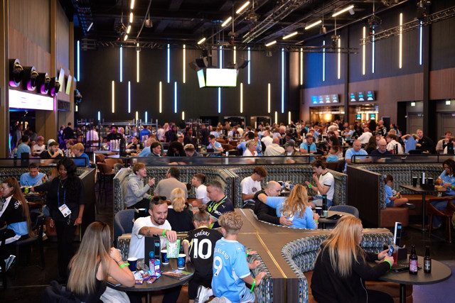 A bustling sports bar filled with Manchester City fans wearing jerseys and enjoying drinks and food around tables. Bright lighting and modern decor create a lively atmosphere.