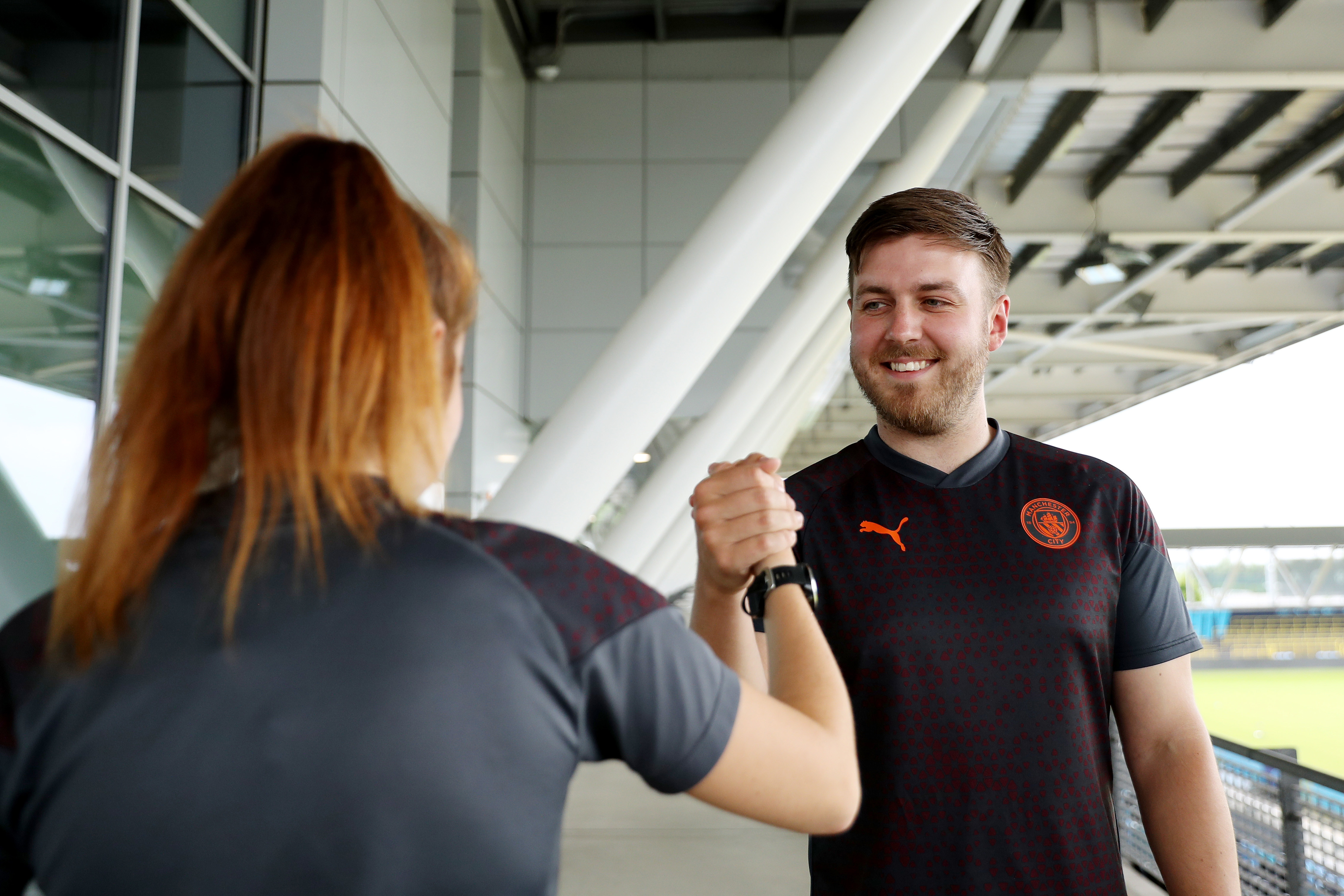 Two people are performing a handshake in a stadium setting, one wearing a Manchester City shirt.