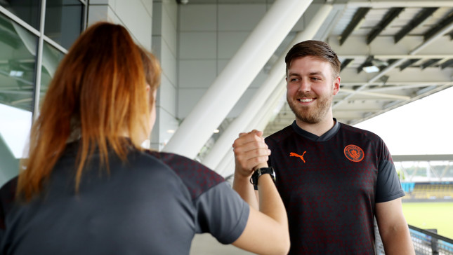 Two people are performing a handshake in a stadium setting, one wearing a Manchester City shirt.