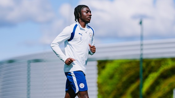 A Manchester City player in training gear jogging outdoors with a cloudy sky in the background.