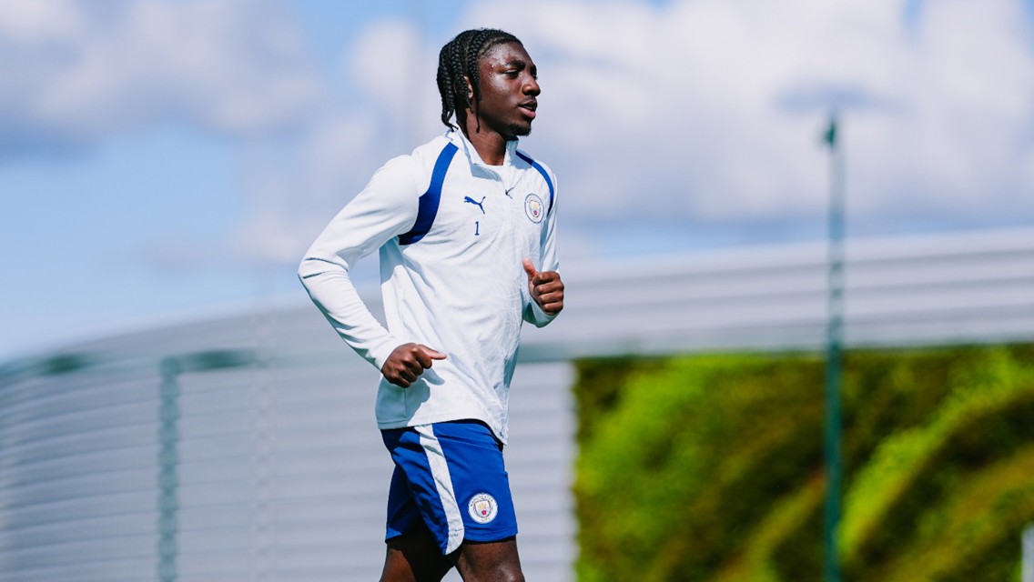A Manchester City player in training gear jogging outdoors with a cloudy sky in the background.