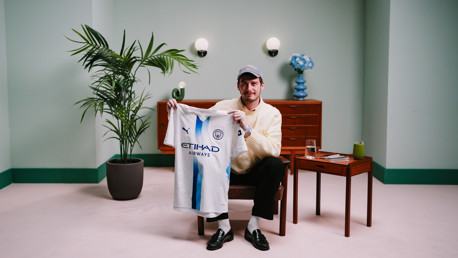 A person holding a Manchester City jersey with 'Etihad Airways' and Puma logos, sitting in a decorated room with plants and furniture.