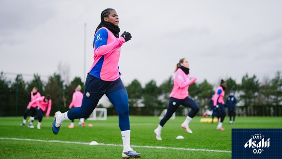 Women's football team training session in pink training bibs outdoors. Players are jogging on a green field with cones. Manchester City crest visible on uniforms.