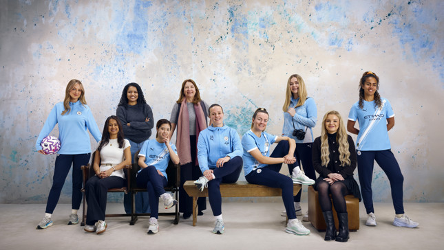 A group of people wearing Manchester City apparel posing for a photo. The background features a speckled blue and white wall.