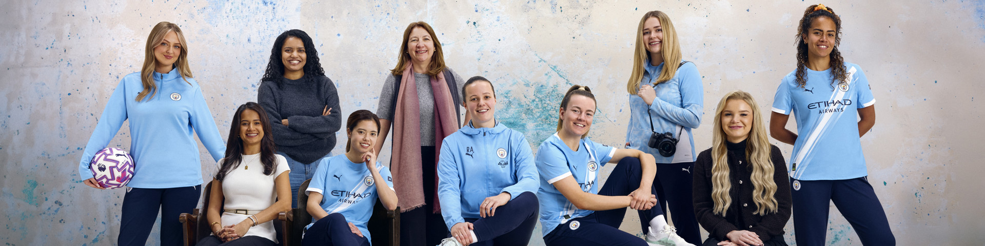 A group of people wearing Manchester City apparel posing for a photo. The background features a speckled blue and white wall.