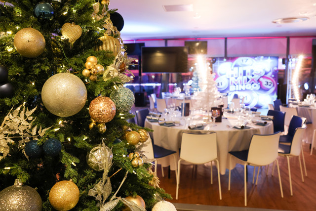 A decorated Christmas tree adorned with ornaments is prominently placed in the foreground. In the background, there is a festively arranged dining area set up for an event, with tables and chairs.