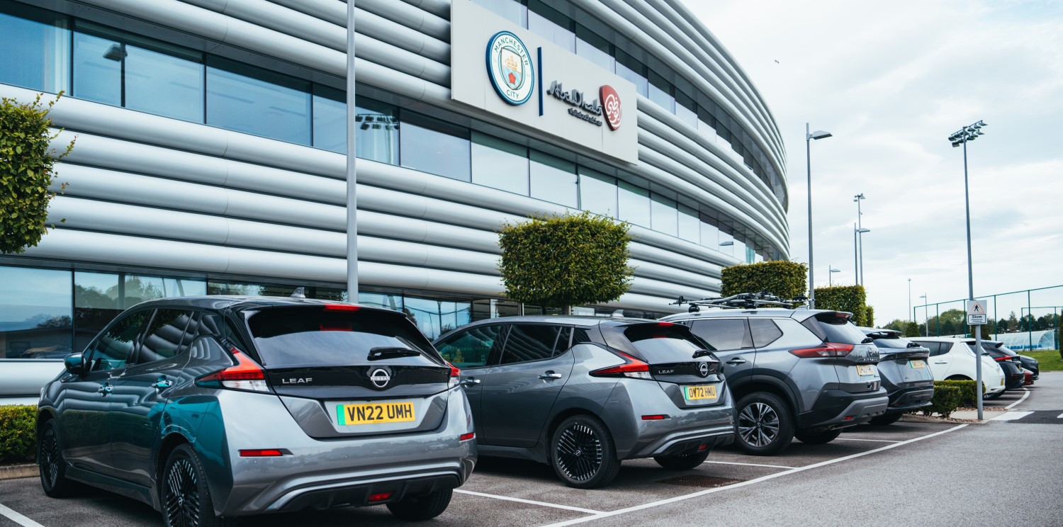 Image of multiple cars, including Nissan models, parked outside the Manchester City stadium with visible Manchester City and Abu Dhabi logos.