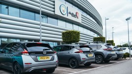 Image of multiple cars, including Nissan models, parked outside the Manchester City stadium with visible Manchester City and Abu Dhabi logos.