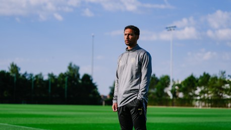 Individual in sports attire standing on a field with a clear sky. The attire has the New York City FC logo.