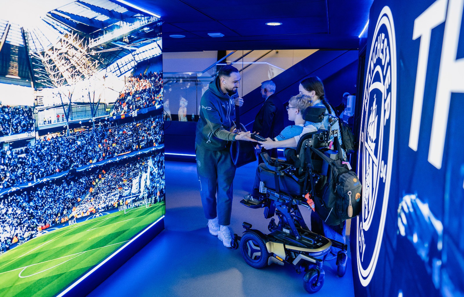 A person in a wheelchair is meeting someone in a dimly lit area, possibly related to Manchester City, as suggested by the logo visible on the wall.