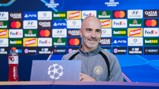 A man in a Manchester City jacket smiles at a press conference table. The backdrop displays UEFA Champions League and sponsor logos. A Gatorade bottle is visible on the table.