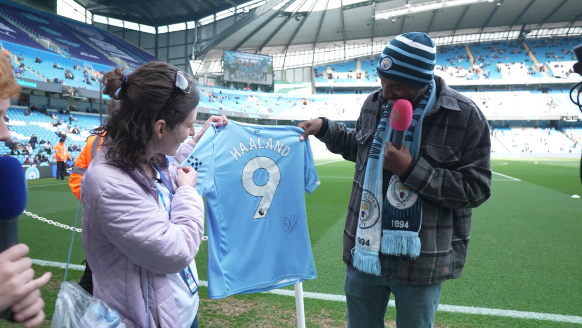 Two people at Manchester City's stadium hold a signed jersey with 'Haaland' and the number 9 printed on it. A microphone and a scarf are visible.