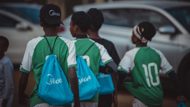 A group of young football players wearing green and white jerseys with numbers on the back, blue drawstring bags, and black caps, walking together near cars.