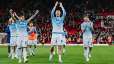 Manchester City players celebrating on the field, wearing their home kit, with fans clapping in the background at a stadium.