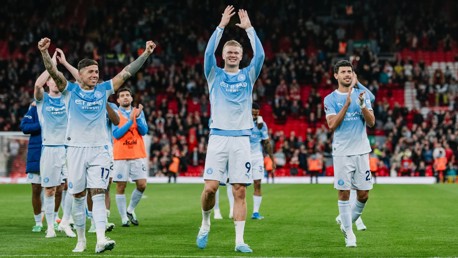 Manchester City players celebrating on the field, wearing their home kit, with fans clapping in the background at a stadium.