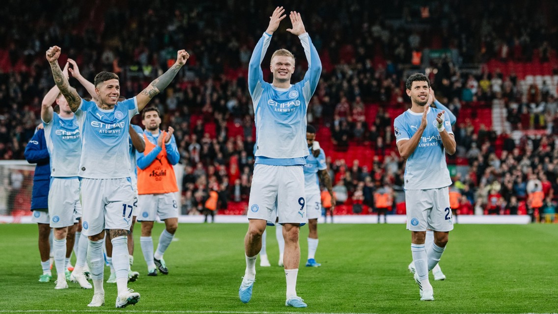 Manchester City players celebrating on the field, wearing their home kit, with fans clapping in the background at a stadium.