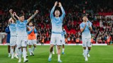 Manchester City players celebrating on the field, wearing their home kit, with fans clapping in the background at a stadium.