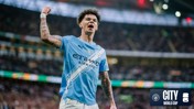 A Manchester City player in a blue jersey celebrates with a raised fist in a stadium. The jersey displays Etihad Airways as the sponsor.