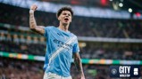 A Manchester City player in a blue jersey celebrates with a raised fist in a stadium. The jersey displays Etihad Airways as the sponsor.