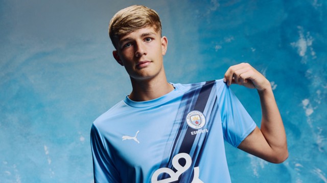 A person holding the Manchester City Esports Puma jersey showing the club's logo on the chest against a blue gradient background.