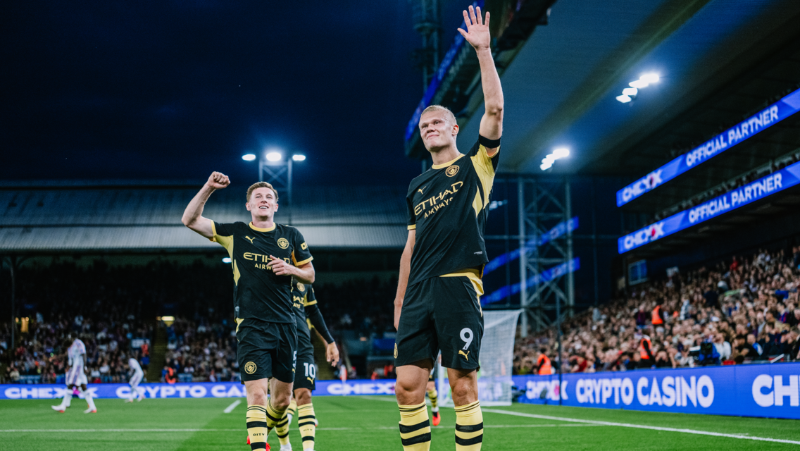 Two Manchester City players celebrating on the field, one waving to the crowd, wearing black and yellow kit. Stadium lights illuminate the scene with cheering fans in the background.
