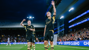 Two Manchester City players celebrating on the field, one waving to the crowd, wearing black and yellow kit. Stadium lights illuminate the scene with cheering fans in the background.
