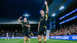 Two Manchester City players celebrating on the field, one waving to the crowd, wearing black and yellow kit. Stadium lights illuminate the scene with cheering fans in the background.