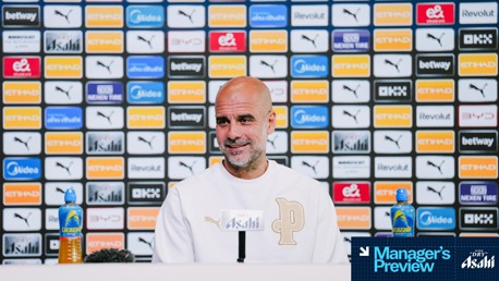 A person with blurred face is seated at a press conference with Lucozade bottles. Background displays various sponsor logos like Etihad, Puma, and Asahi. Text reads 'Manager's Preview'.