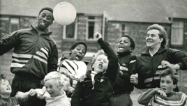 Group of children and adults playing outdoors with a ball. The setting appears to be a playground or sports field.