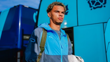 A man is standing in front of a team bus, wearing a blue and gray jacket with a Puma logo, carrying a bag.