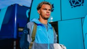 A man is standing in front of a team bus, wearing a blue and gray jacket with a Puma logo, carrying a bag.