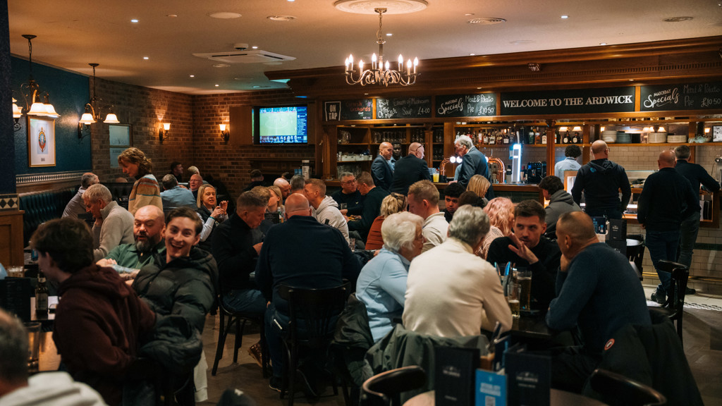 An interior view of a pub named 'The Ardwick' with numerous patrons sitting at tables, socializing and dining. The pub has brick walls, a chandelier, and menu boards with specials written on them.