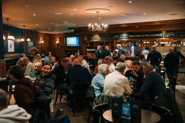 An interior view of a pub named 'The Ardwick' with numerous patrons sitting at tables, socializing and dining. The pub has brick walls, a chandelier, and menu boards with specials written on them.