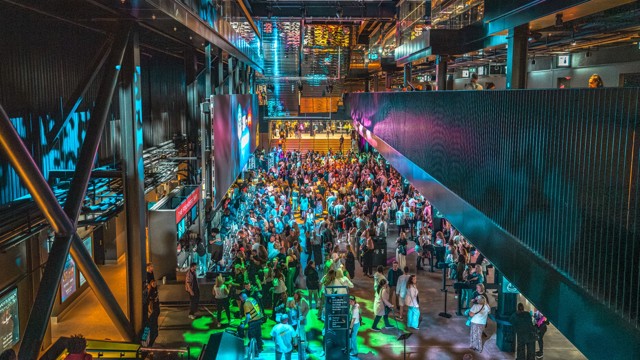 A large crowd of people gathered in a modern venue with colorful lighting and multiple levels.