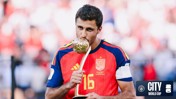 A male football player wearing a Spain jersey with the number 16 is kissing a trophy. There is a City at the World Cup logo on the bottom right.