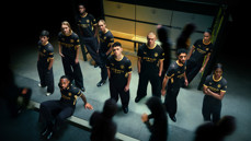 Group of individuals wearing Manchester City 2023 black and yellow kit with Etihad Airways sponsor, standing and sitting indoors.